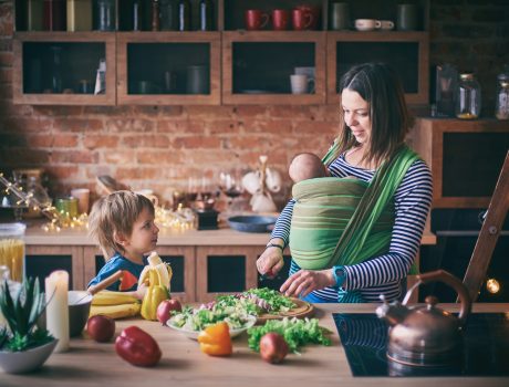 Happy young family, beautiful mother with two children, adorable preschool boy and baby in sling cooking together in a sunny kitchen.